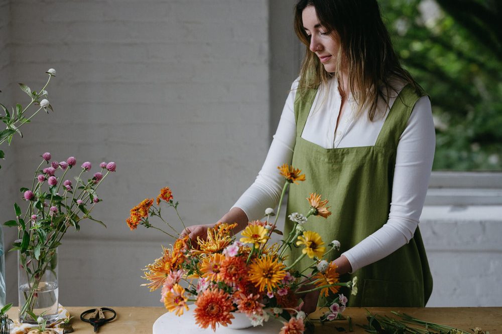 White woman in green criss cross apron arranges orange and yellow flowers