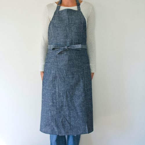 person modeling a split leg apron in linen. they are wearing a white shirt and jeans and standing in front of a white wall. the apron is denim.