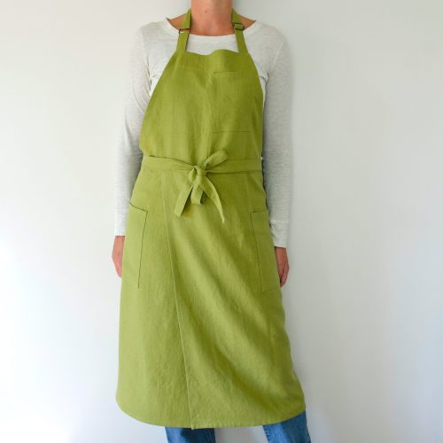 person modeling a split leg apron in linen. they are wearing a white shirt and jeans and standing in front of a white wall. the apron is green.