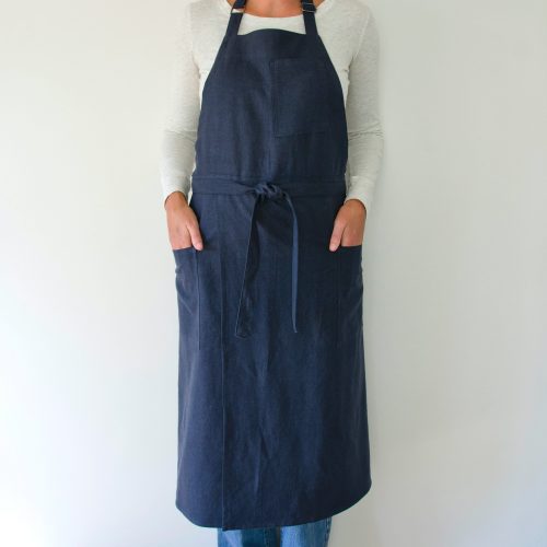 person modeling a split leg apron in linen. they are wearing a white shirt and jeans and standing in front of a white wall. the apron is blue.